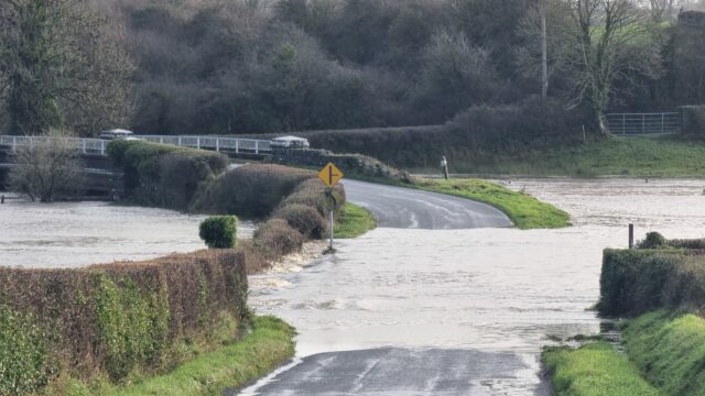 A road through a field and towards a river bridge is deeply submerged under fast-flowing water.