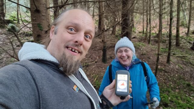 Dan and his mother smile for the camera in a forest, holding a GPSr between them.