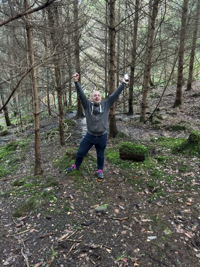 Dan raises his hands in a victory pose in a forest.