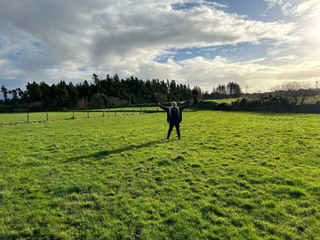 Dan raises his hands - one of them holding a GPS receiver - in the air in a victory pose, in a green pasture with a forest in the background.