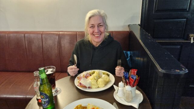 A woman sits in front of a plate loaded with food in a pub setting, holding a knife and fork.