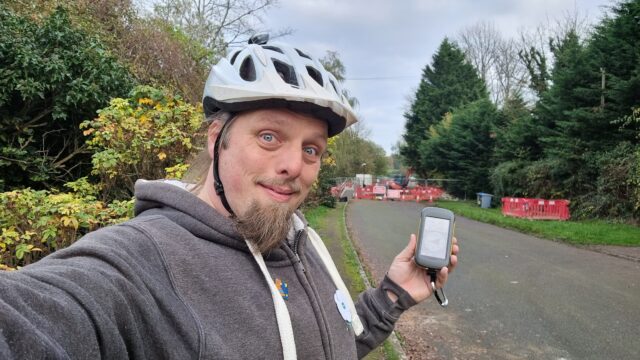 Dan smiling, wearing a cycle helmet, holding a GPSr by the side of a narrow road, with roadworks in the background.