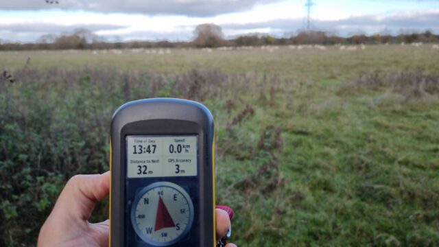 GPS receiver in front of a field. The compass points deeper into the field and the screen reports that the destination is 32 metres away. Sheep are (barely) visible in the field, in the distance.