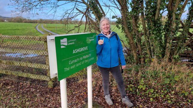 A smiling woman with white hair, wearing spectacles atop her head, stands next to a sign that reads 'Ashgrove: The wet land of the curses' in English and Irish. She holds a small tube-shaped geocache.