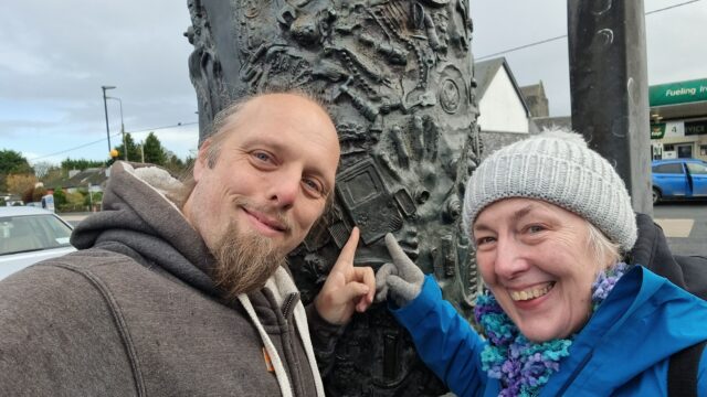 A cylindrical metal sculpture stands upright on a spindle, in a manner slightly reminiscient of a kebab. It's decorated with hundreds of small contemporary handheld objects such as spanners, rings, spoons, and chess pieces. Dan and his mother Doreen stand in front, both pointing to a Nintendo Game Boy.