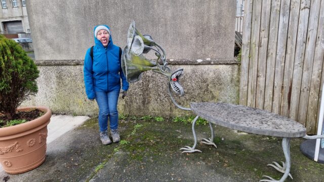 A sculpture (called 'gossip') is a bench with birdlike feet and a head comprised of a mouth with a long tongue and several listening tubes. Alongside it, Dan's mother - a woman wearing a grey bobble hat and a blue raincoat - attempts to mime the same mouth shape.