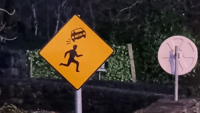 Photograph showing a road sign; it's yellow, diamond-shaped, and depicts in black the silhouette of a person running from left to right. Above them is the silhouette of a car, much smaller than them and twisted anticlockwise by about 20 degrees. Impact/movement marks eminate from the lower of the car's wheels, as if it's a thrown object that's just bounced off the head of the runner.