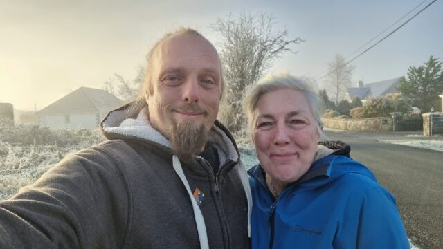 Dan and his mother, smiling, in frozen countryside, with fog behind.