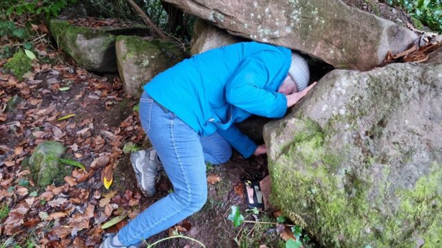 A woman in a bobble hat and a blue raincoat crawls into a gap between two boulders.