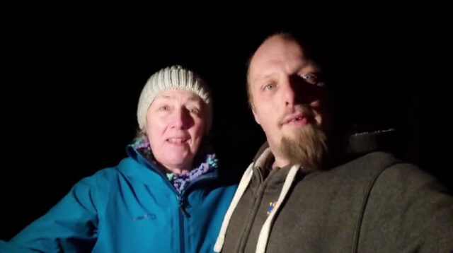 Dan and his mum, lit by phone-light, stand in a dark forest.