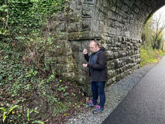 Dan, standing by a bridge, shakes a logbook out of a tubular geocache.