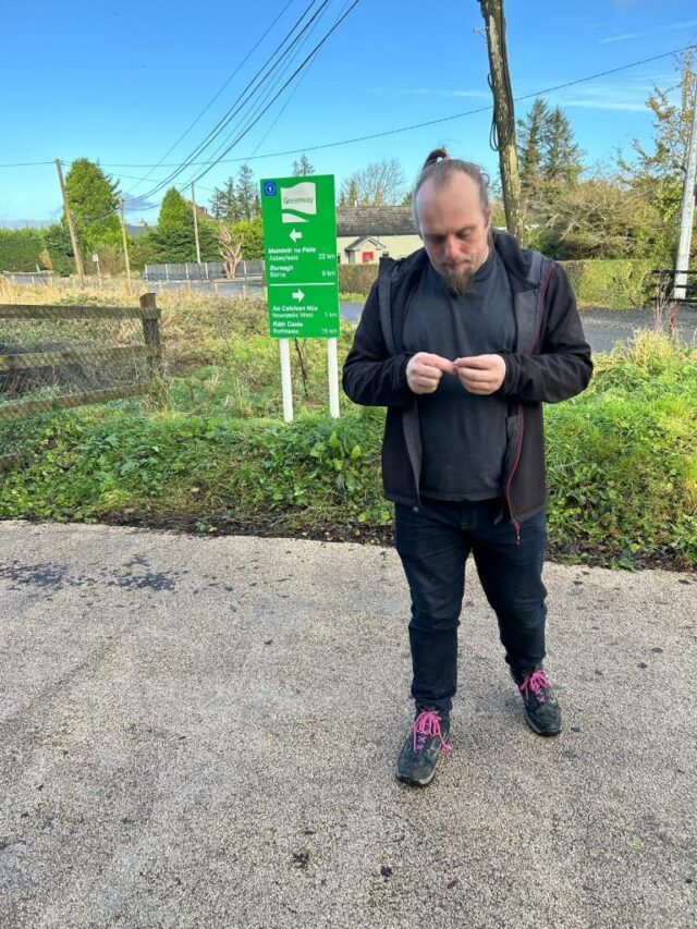 Dan, standing by a "Greenway" sign, unrolls a geocaching logbook.