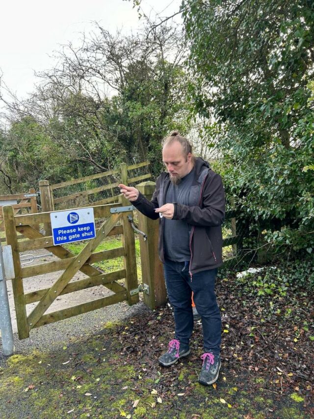 Dan standing by a gate, reading a geocaching logbook.