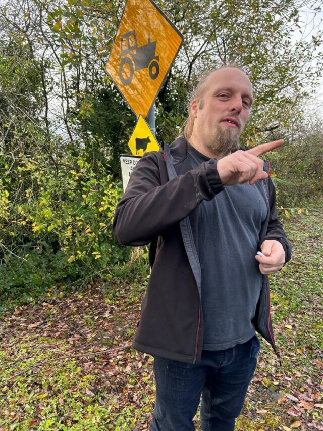 Dan, with his finger sticking out, stands in front of 'cow crossing' and 'tractor crossing' signs.