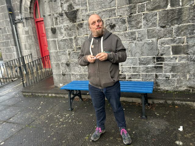 Dan stands in front of a blue bench, in the rain, in front of a stone wall.