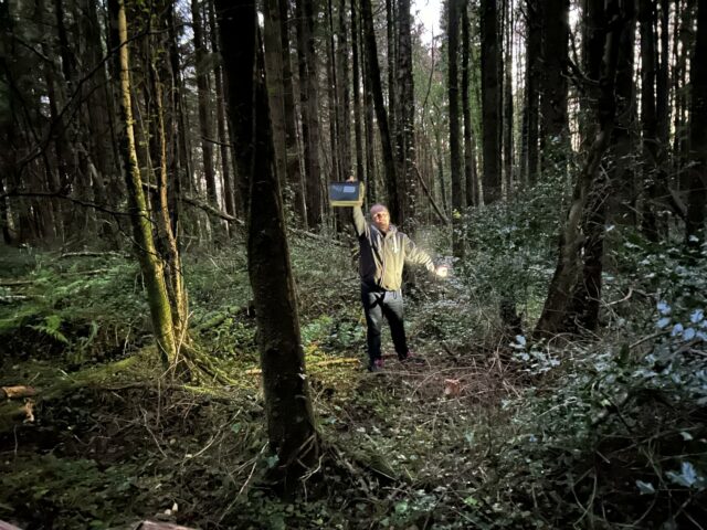 Dan, in a dense forest at twilight, holds aloft a large ammo can. His other hand holds his phone, used as a flashlight.