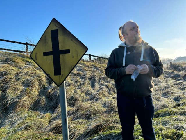 Dan smiles as he opens a zip-lock bag containing a geocaching logbook, alongside a diamond-shaped Irish 'crossroads' road sign.