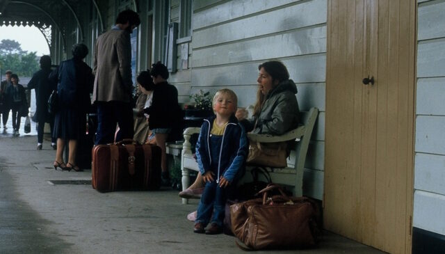 Dan, aged ~4, stands on a railway station platform alongside his mother, yawning, on a bench. It is overcast and drizzly.