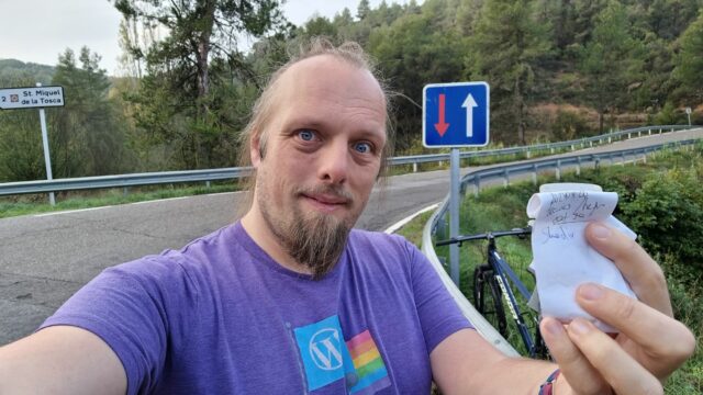 Dan, holding a small geocache logbook. A bike and a road bridge can be seen in the background.