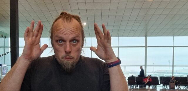 Dan, standing in an airport departure lounge, mimes "mind blown" to the camera.