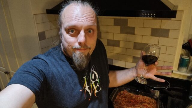 Dan, in a kitchen with some kind of tomato sauce on a hob behind him, holds a glass of red wine and shrugs at the camera.