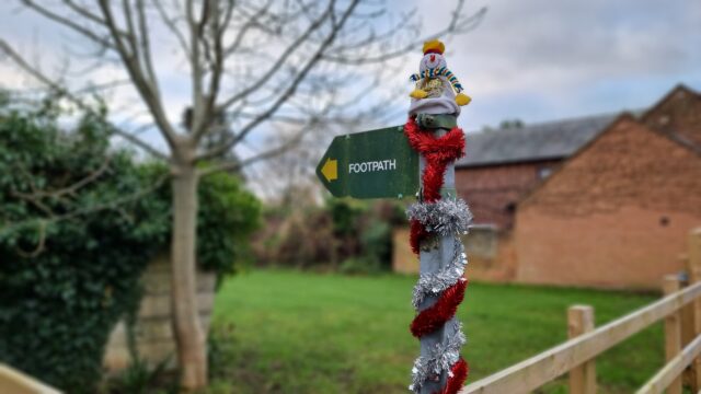 Finger post footpath sign adorned with a toy snowman and wrapped in a spiral of red and silver tinsel.