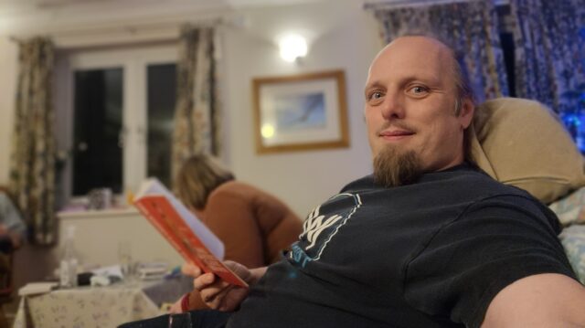 Dan sits by firelight reading a red-spined book.