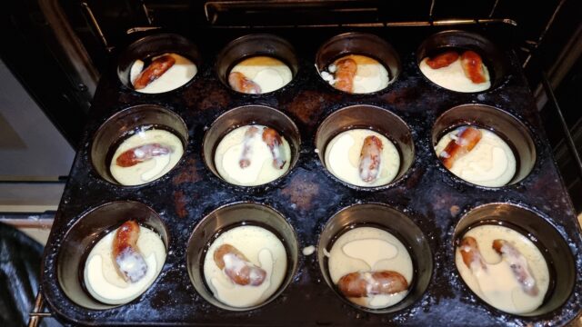 Cupcake-sized Yorkshire pudding batter cups, each with a cocktail sausage or two inside, being inserted into an oven.