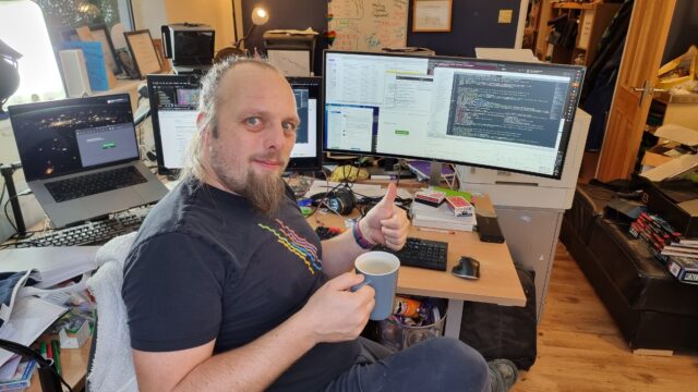 Dan, a white man with a goatee-style beard and long hair tied back, wearing a black t-shirt with an abstract pride rainbow, holds a mug of coffee in one hand and gives a thumbs-up with the other. Behind him, three computer screens show a mixture of code, source control, chat, email, and web browser windows. His desk is cluttered.