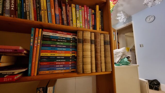 A bookcase in a child's bedroom, with Christmas decorations visible in the background. Several very old looking books stand conspicuously on the shelf.