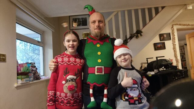 Three people in a decorated hallway, wearing Christmas jumpers and headgear. Dan, in the centre, is wearing a jumper designed to make him look like a tiny elf, and a matching hat. To his right, a girl wears a jumper showing Rudolf, and a pair of spring-mounted reindeer deely-boppers. To his left, a boy with his eyes closed throws a thumbs-up: he's wearing a jumper with a pixel-art picture of Santa, and a wooly Santa hat.