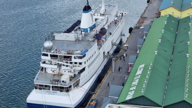 White and blue passenger ship docked alongside a building whose roof reads 'Welcome to Port of Spain'.