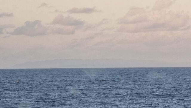 Across the sea, a rugged island begins to emerge over the horizon, through distant clouds.