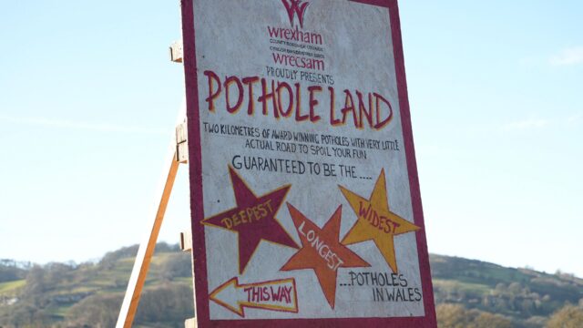 Humorous handmade road sign welcoming visitors to Wrexham as 'Pothole Land', stating that there are 'two kilometres of award-winning potholes with very little actual road to spoil your fun' and guaranteeing 'the deepest, longest, vs widest potholes in Wales'.