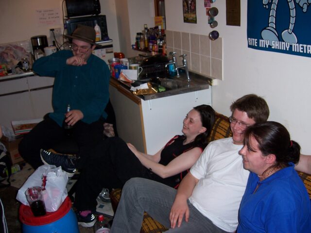 Four young people, smiling in laughing, sit in a cluttered and messy flat.