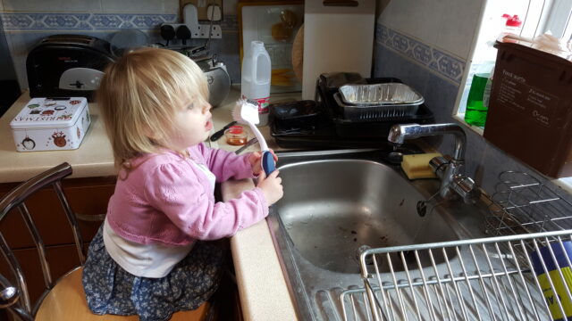 A toddler with long blonde hair, wearing a pink cardigan, sits on a tall stool in front of a kitchen sink, holding a long-handled scrubbing brush.