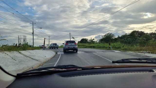 View through a windscreen showing cars ahead going over a speed bump and winding along a road between tropical shrubbery, with power lines overhead.