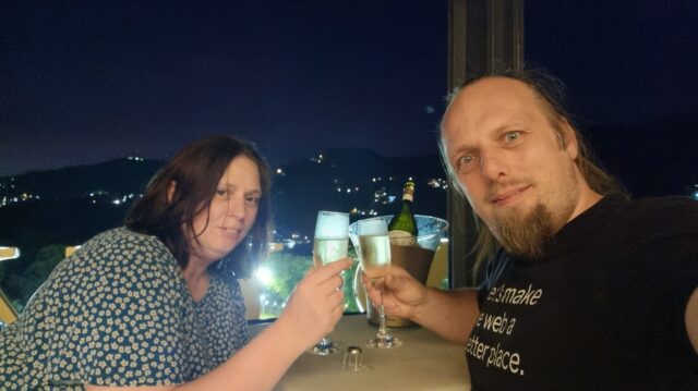 Dan and Ruth clink champagne flutes under a dark sky, with city lights visible below them.