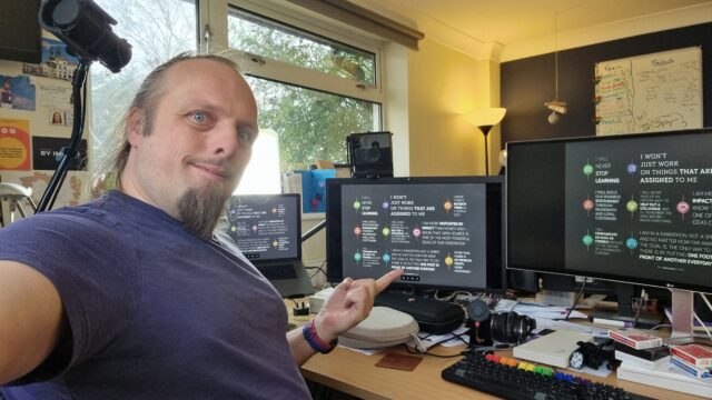 Dan, wearing a purple t-shirt, looks at the camera while pointing at the centre of three computer screens which share a cluttered desk, each of which shows a stylised image version of the Automattic Creed.