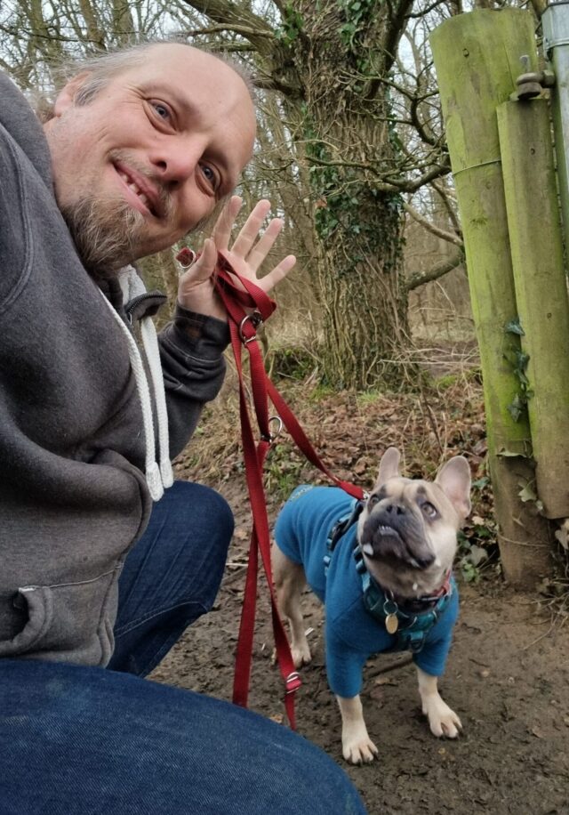 Dan, wearing a grey jacket at the edge of a wet and wintry forest, waves to the camera with a hand that also holds a dog lead, the other end of which is connected to a French Bulldog wearing a teal jumper.