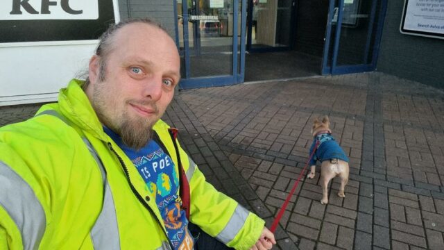 Dan, wearing a high-vis jacker, sits at a bench outside a motorway service station building. His dog, a small French Bulldog, pulls at her lead towards the entrance.