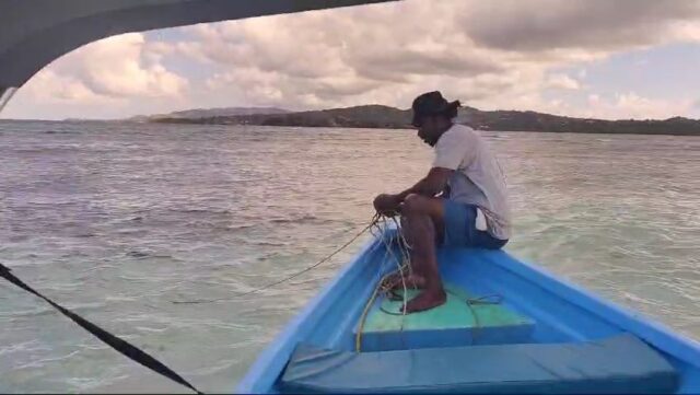 A man sits on the prow of the Cariad, over a sandbar, pulling in her anchor.