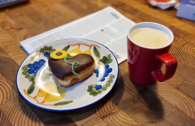 A wooden kitchen surface containing a red mug full of freshly-brewed coffee alongside a plate painted with fruits on which sits a heart-shaped doughnut, topped with chocolate and decorated with an iced motif of a sunflower. Beneath the plate, out-of-focus, are the pages of a news periodical.