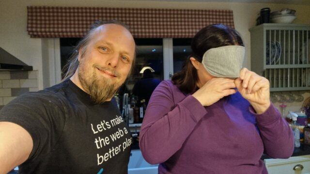 Dan, wearing a black t-shirt, smiles as he takes a selfie. Alongside him Ruth, wearing a purple jumper, adjusts a grey blindfold to cover her eyes.