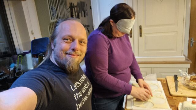 Dan takes a selfie showing himself, smiling, and Ruth, wearing a blindfold and balling up pastry on a wooden worksurface.