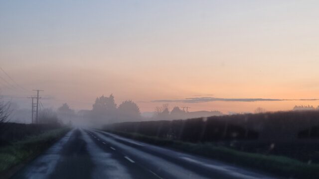 A wet rural road, lit by the sun's first light as it rises ahead, it's flanked by fields full of freezing fog.