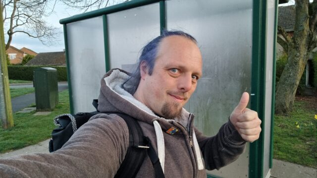 Dan, a white man with a trimmed goatee beard and dyed-blue hair tied back in a ponytail, stands alongside a suburban bus shelter and throws a thumbs-up sign. He's carrying a backpack.