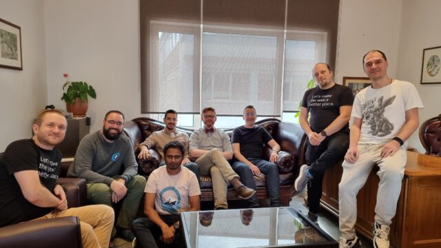 A group of men sit on chairs, a sofa, and the edge of a desk in a comfortable large office space.