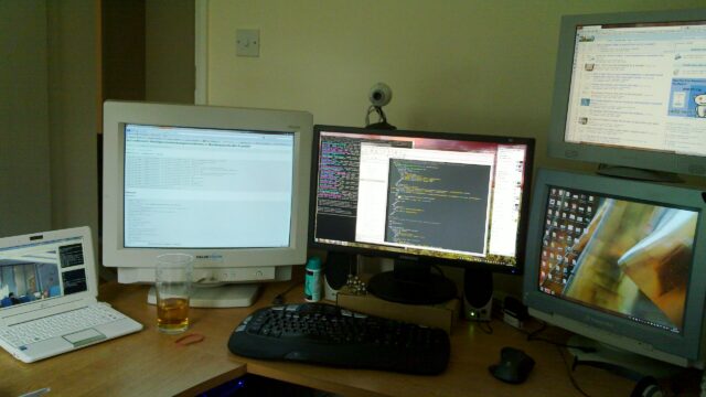 A mixture of flatscreen and CRT monitors, plus a laptop and a webcam, on a desk. The laptop screen shots a view of an office at the "other end" of a webcam connection.