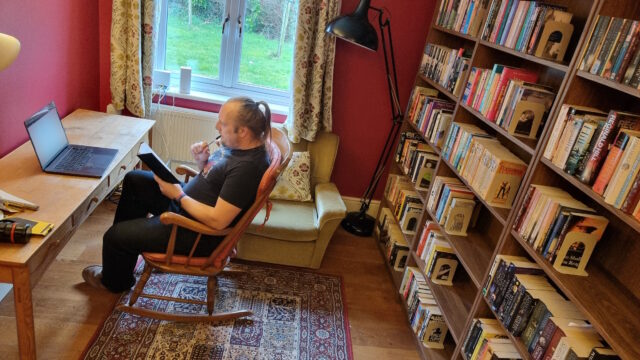 Dan, a white man with a ponytail, wearing a black shirt and jeans, sits in a rocking chair in front of an open laptop at a long desk; he has a notebook in his hand and holds a pen near his lips. He's in a domestic library with deep red walls, balanced-arm lamps, a woven rug on a wooden floor, and the wall behind him entirely covered with floor-to-ceiling bookshelves. A grassy lawn and sapling trees can be seen through a window, bordered by floral curtains and two clumsily-placed WiFi routers that sit on the window ledge. On the corner of the desk lie various hand tools, suggesting that light construction work has recently taken place.